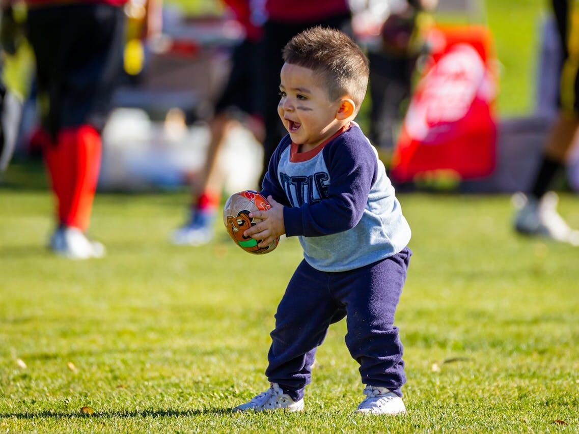 Joyful toddler playing with a football on a sunny day outdoors, dressed in casual clothes.