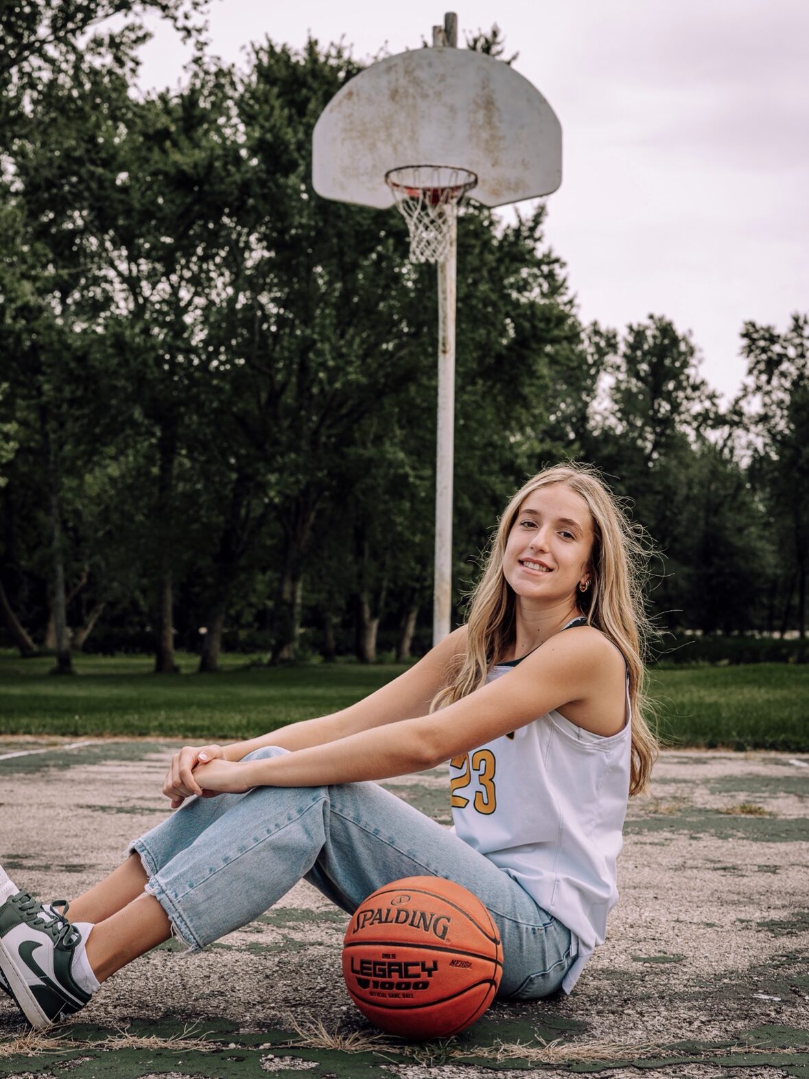 Upload up to 5 photos that you think should be shared as part of the Player Spotlight Min size 800 x 800 pixel IMG_0097 - Ball to Life Smiling young woman sits on a worn outdoor basketball court with a basketball beside her and a hoop in the background.
